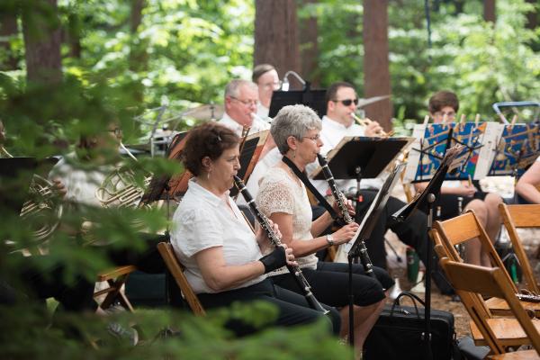 Orchestra members play their instruments in a wooded setting.